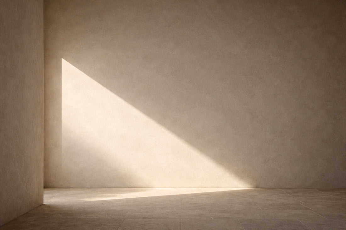 Minimalistische Architekturfläche aus hellem Kalkputz und Steinboden. Seitliches Vormittagslicht bildet ein klar begrenztes, trapezförmiges Lichtfeld auf der Wand, umgeben von weichen Schatten.