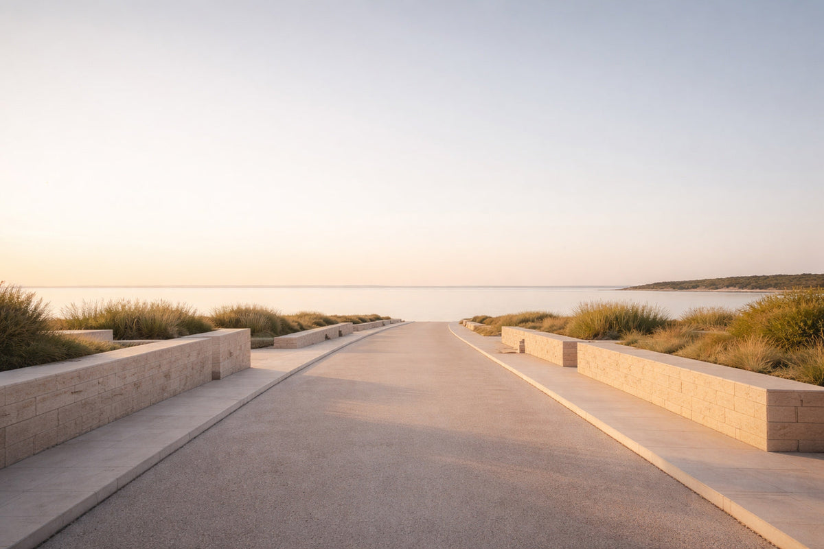 Weiter, leerer Straßenraum aus hellem Asphalt führt ohne Ziel auf einen ruhigen Horizont zu. Früher Abend, noch helles Licht ohne Dramaturgie. Sanfte Kontraste, klare Linien, niedrige Steinbegrenzungen,