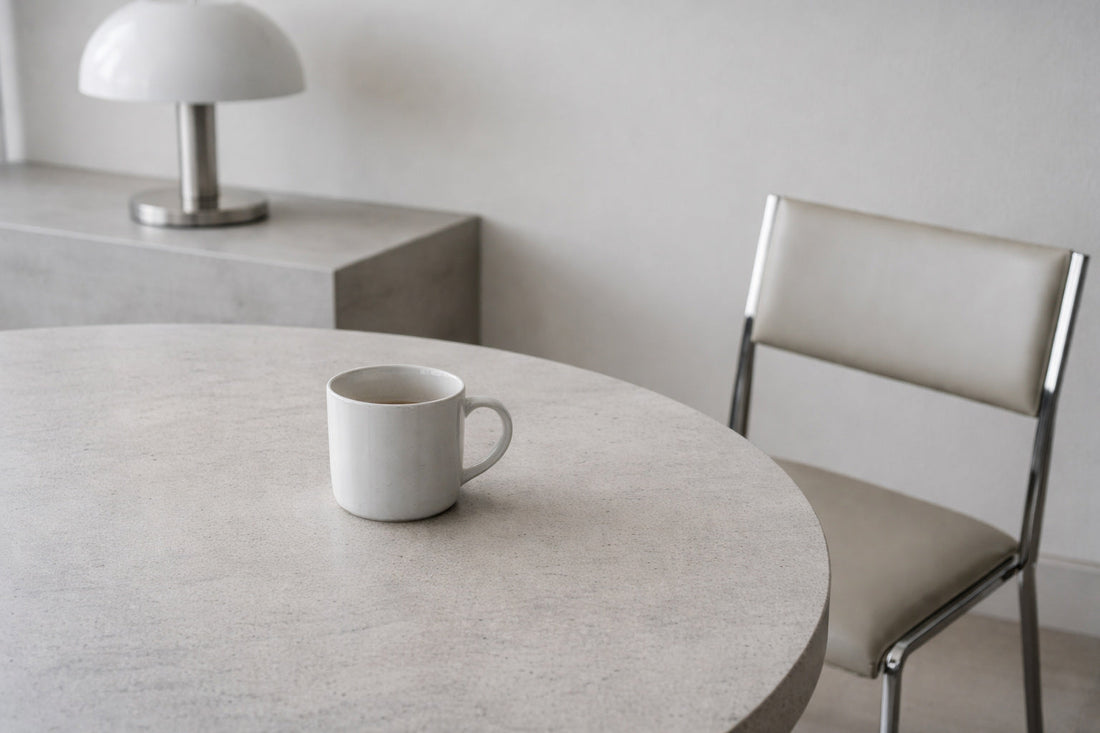 Minimalistischer Innenraum am Vormittag mit ruhigem, sachlichem Licht. Ein einzelner weißer Keramikbecher steht auf einem hellen Stein- oder Betontisch. Materialität und leichte Gebrauchsspuren sind sichtbar.