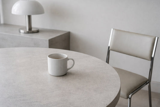 Minimalistischer Innenraum am Vormittag mit ruhigem, sachlichem Licht. Ein einzelner weißer Keramikbecher steht auf einem hellen Stein- oder Betontisch. Materialität und leichte Gebrauchsspuren sind sichtbar.