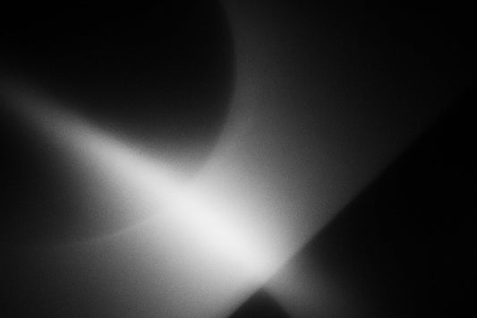 Ein abstraktes Schwarz-Weiß-Bild, in dem ein schräger Lichtstrahl durch dunkle Flächen bricht. Weiche Übergänge, kreisförmige Schatten und eine diagonale helle Linie erzeugen eine stille