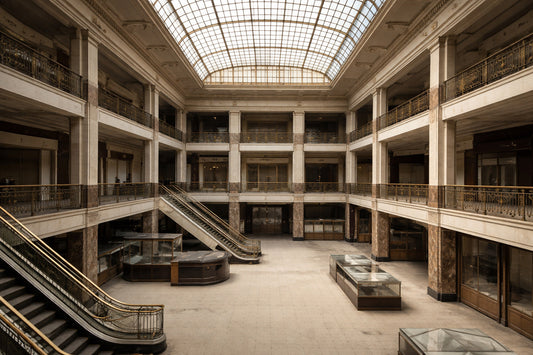 Leere Kaufhaushalle aus den frühen 1920er-Jahren mit mehreren Ebenen und offenen Galerien. Sachliches Mittagslicht fällt flächig in den Raum, gealterte Materialien und architektonische Details sind sichtbar.