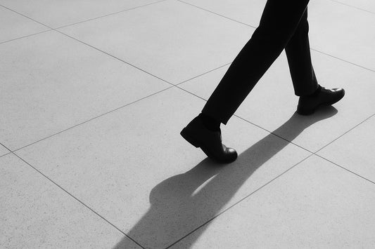 Diagonal verlaufender Schatten eines gehenden Menschen auf hellem Steinboden; nur Beine und Bewegung sichtbar, reduziertes Schwarz-Weiß, ruhig und offen ohne Ziel.