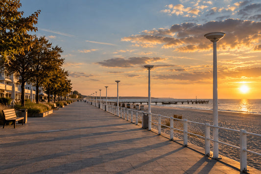 Menschenleere Strandpromenade in der goldenen Abendsonne, mit Bäumen, Laternen und einer weißen Reling zum Meer hin, im Hintergrund ein Steg im warmen Herbstlicht.