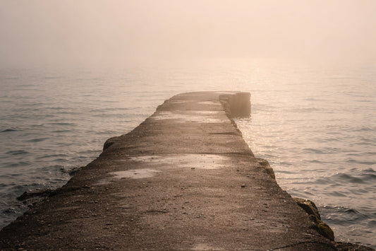Schmaler, verwitterter Betonsteg, der in ruhigem Wasser im Morgennebel verschwindet; warmes, diffuses Licht, keine Personen, Gefühl von Weitergehen ohne Ziel oder Abschluss.