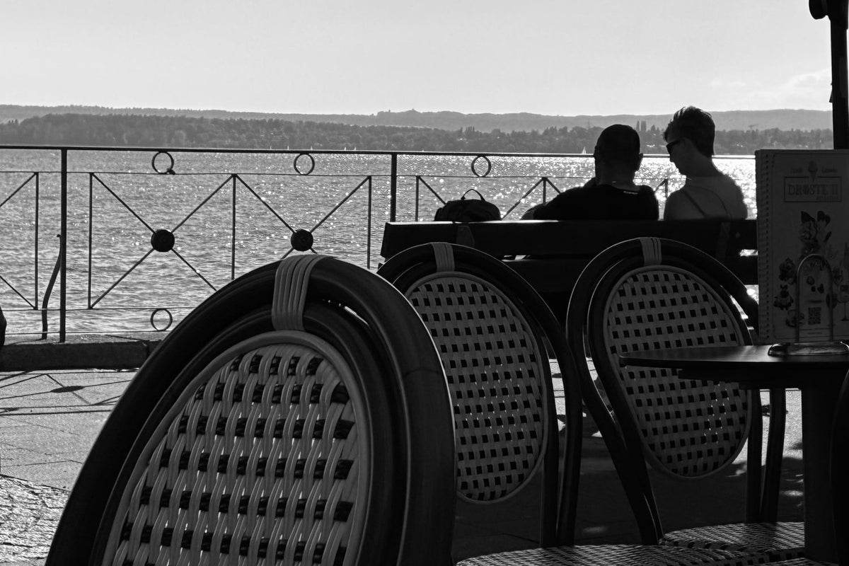 Rattan-Stühle im Gegenlicht auf einer Terrasse am Bodensee, zwei Silhouetten am Tisch im Hintergrund, schimmerndes Wasser
