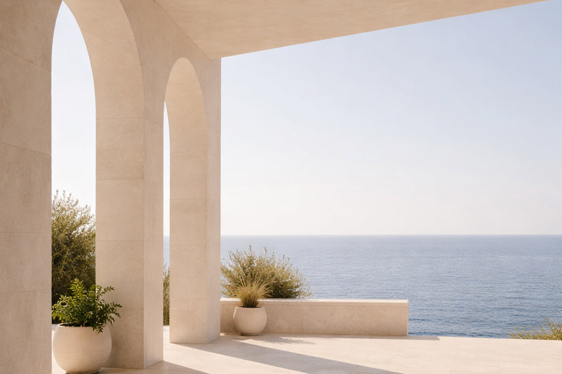 Helle mediterrane Terrasse aus weißem Stein mit klaren Bögen und weitem Blick auf das ruhige Meer; warmes Nachmittagslicht, lange Schatten, offene Architektur, stille Weite ohne Menschen.