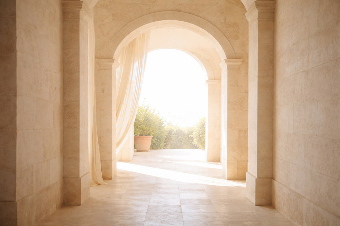 Sanftes mediterranes Licht fällt durch einen hohen Steinbogen aus hellem Naturstein; weiße Marmorsäulen, ein halbtransparenter Vorhang im Wind und eine ruhige Terrasse im Hintergrund