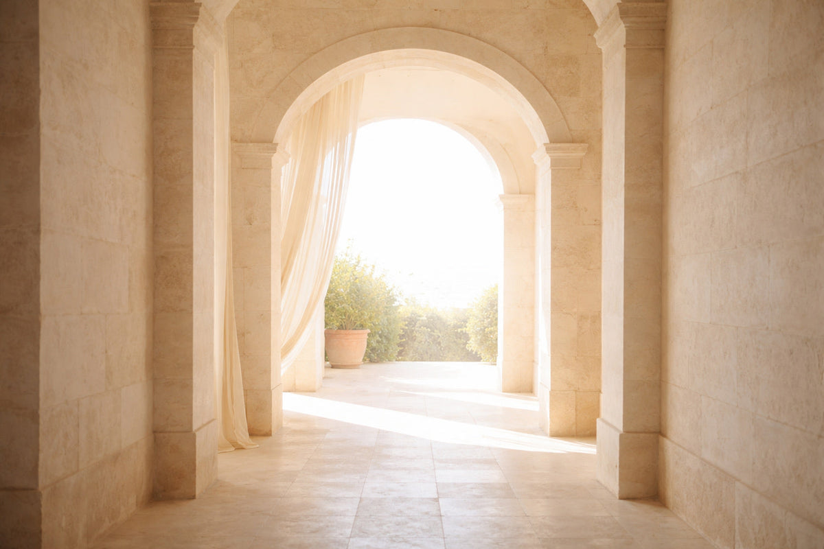 Sanftes mediterranes Licht fällt durch einen hohen Steinbogen aus hellem Naturstein; weiße Marmorsäulen, ein halbtransparenter Vorhang im Wind und eine ruhige Terrasse im Hintergrund