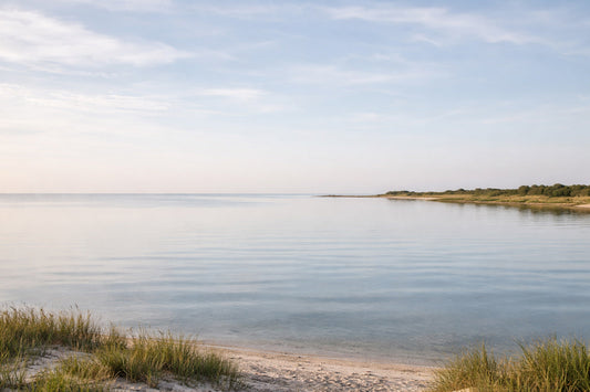 Ruhige Küstenlandschaft mit flachem Horizont, stillem Wasser und hellem Himmel. Das Wasser liegt fast unbewegt, die Küstenlinie zieht sich leise in die Ferne. Keine Menschen, keine Boote,