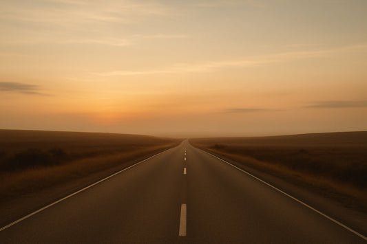 Lange, gerade Landstraße im warmen Abendlicht, die in einen goldenen Horizont führt. Weiche Schatten liegen über den Feldern, der Himmel zeigt sanfte Gold- und Orangetöne. Ruhige, weite Szene eines stillen Heimwegs.