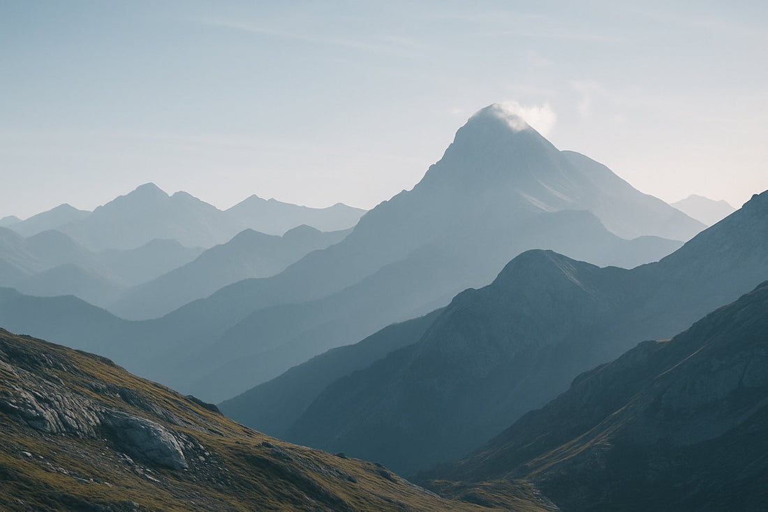 Weite, klare Berglandschaft der Pyrenäen mit gestaffelten blaugrauen Höhenzügen und einem wolkenumspielten Gipfel im Licht. Raues Gestein im Vordergrund, ruhige, lichte Atmosphäre.