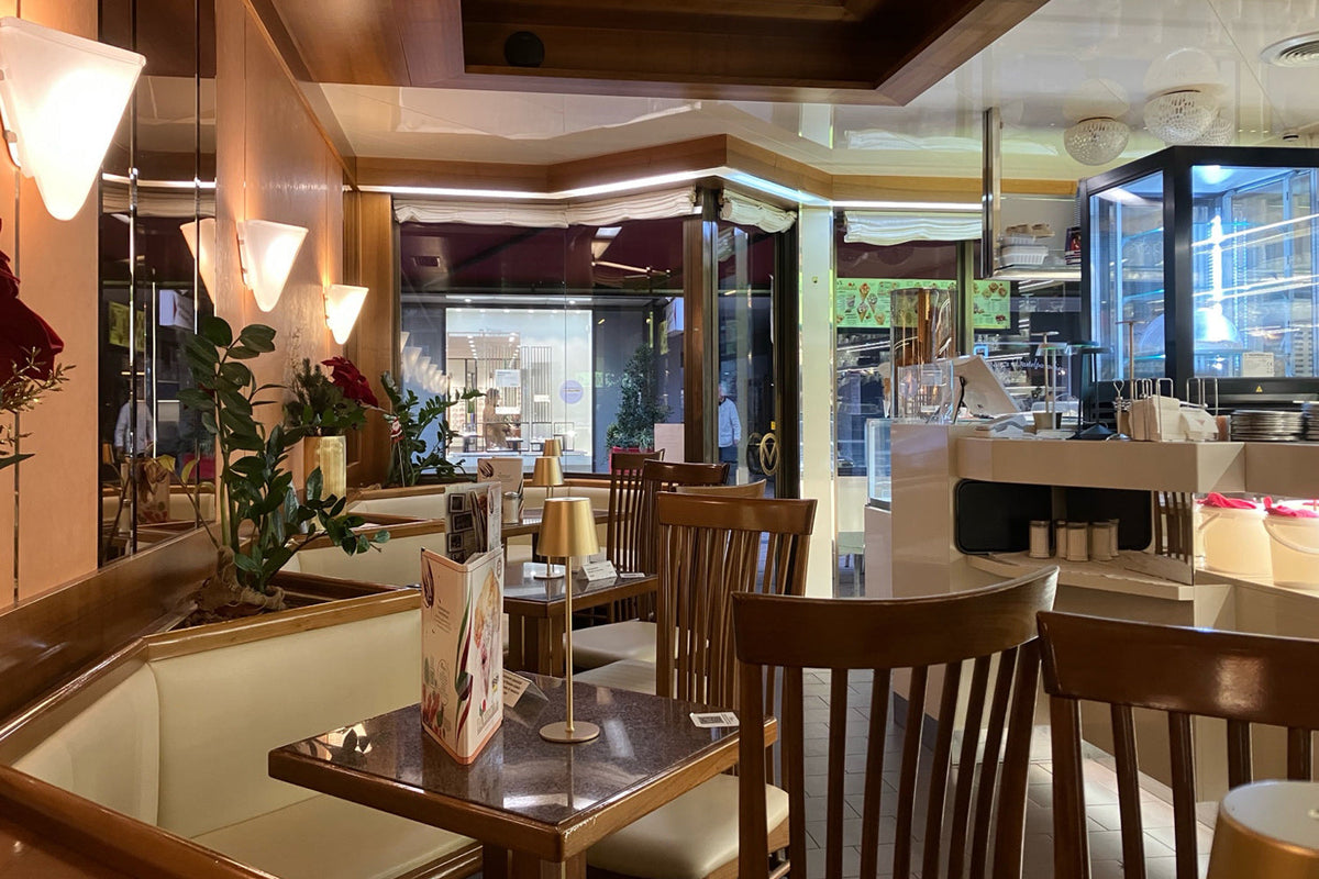 Innenraum eines italienischen Eiscafés am Morgen: kleine Tische, Holzstühle, warmes Licht, ruhige Atmosphäre, ein gedeckter Tisch mit Lampe und Menükarte, Blick nach draußen auf den beginnenden Tag.